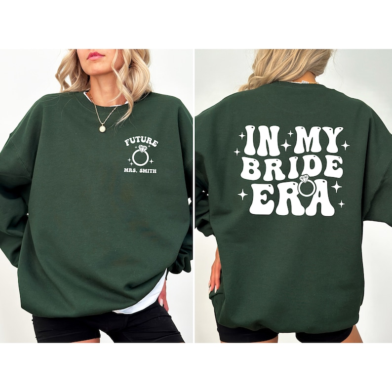 May include: A dark green sweatshirt with white text that says "Future Mrs. Smith" on the front and "In My Bride Era" on the back. The text is in a retro font with a ring and stars.