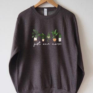 Puede incluir: Una sudadera gris oscuro con un gr&aacute;fico blanco de plantas en macetas y el texto "just one more".