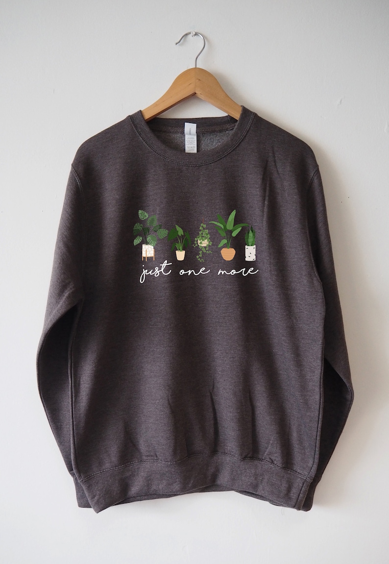 Puede incluir: Una sudadera gris oscuro con un gr&aacute;fico blanco de plantas en macetas y el texto "just one more".