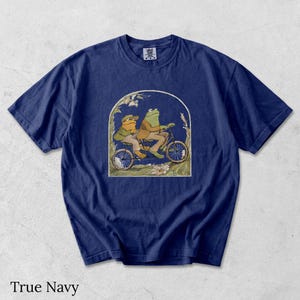 May include: A true navy blue short-sleeve t-shirt featuring a graphic print of two anthropomorphic frogs riding a tandem bicycle. The illustration is framed within an arch and depicts the frogs wearing brown jackets and hats in a grassy setting.