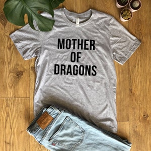 May include: A heather gray t-shirt with the text "MOTHER OF DRAGONS" in black. The shirt is laid on a wooden surface next to a pair of light blue jeans. Decorative plants are in the background.