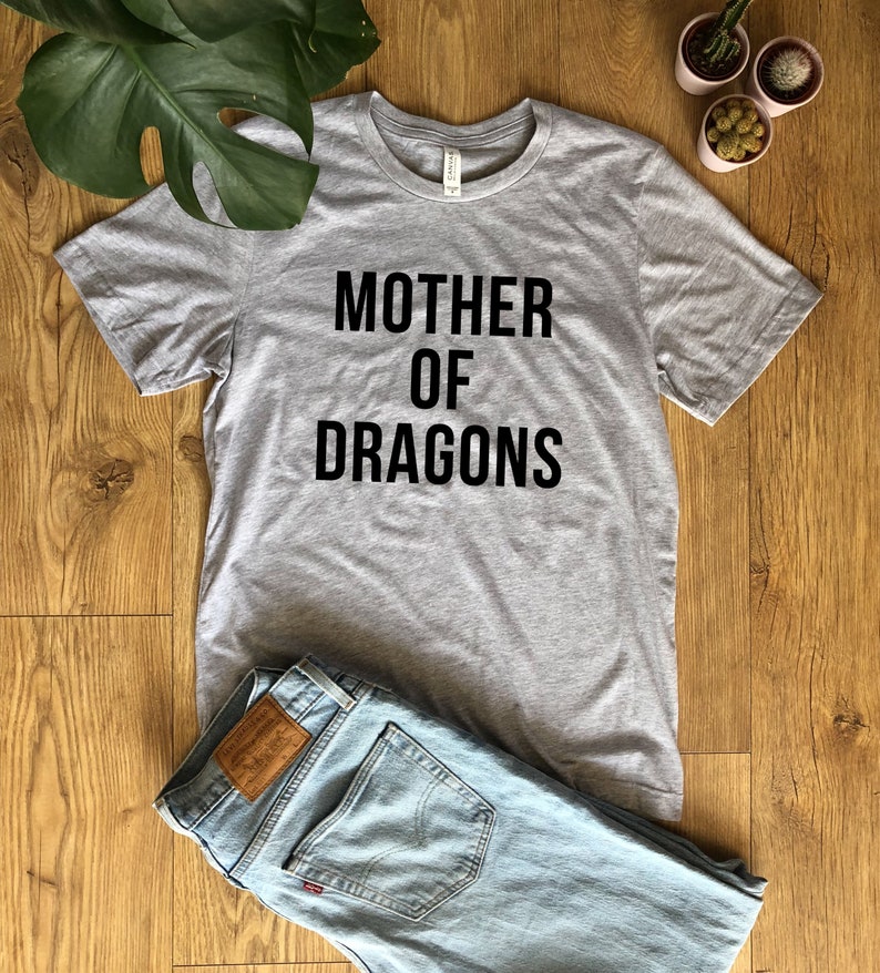 May include: A heather gray t-shirt with the text "MOTHER OF DRAGONS" in black. The shirt is laid on a wooden surface next to a pair of light blue jeans. Decorative plants are in the background.
