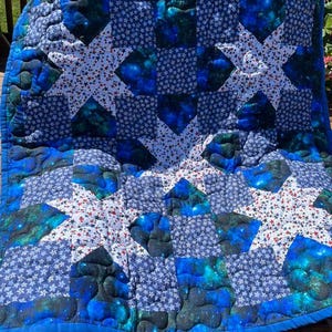 May include: A quilted blanket featuring a star pattern composed of white fabric with small red and blue stars, dark blue fabric with white star prints, and a vibrant blue and green nebula-inspired print with a solid blue fabric border.