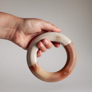May include: A circular ceramic object with a smooth, off-white glazed upper section and a textured, reddish-brown unglazed lower section. A small, stamped maker's mark is visible on the upper white portion of the ring-shaped piece.