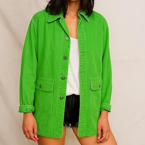 Peut inclure: Une femme porte une veste en jean vert vif avec deux poches avant et un chapeau noir. La veste est boutonnée et portée sur un haut blanc et un short noir. La tenue est décontractée et élégante.