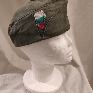 May include: Olive green military-style cap with a small, colorful patch featuring white, green, and red stripes. The cap is displayed on a white mannequin head, showcasing its shape and design. The cap appears to be made of a durable fabric.