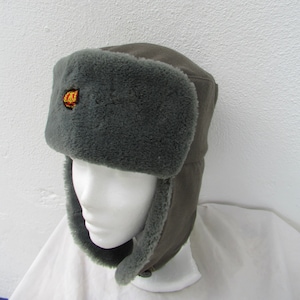 Può includere: Un cappello Ushanka grigio con paraorecchie e un distintivo. Il cappello presenta un esterno grigio scuro e una fodera interna più chiara e soffice. Il distintivo è rosso e oro. Il cappello è esposto su una testa di manichino bianca.