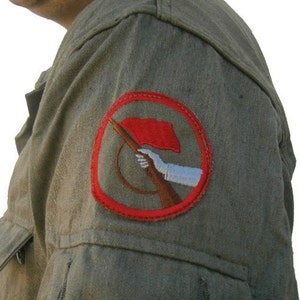 Puede incluir: Chaqueta militar verde oliva con dos bolsillos delanteros y un parche rojo y blanco en la parte superior del brazo. El parche presenta una bandera roja, un rifle y un brazo blanco sosteniendo el rifle.