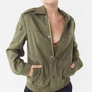 Puede incluir: Chaqueta bomber verde oliva con cuello, cierre de botones y cremallera, y bolsillos. La chaqueta tiene dobladillo y puños acanalados. La modelo lleva un top verde oliva a juego y pantalones negros.