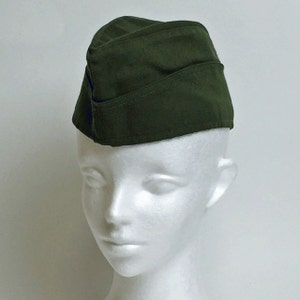 May include: A dark green military-style cap, also known as a garrison cap, is displayed on a white mannequin head. The cap is made of a soft fabric and features a folded design. The cap's color is a deep, muted green, and it is set against a plain white background.