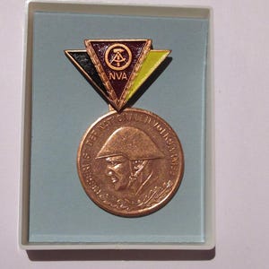 May include: A vintage medal with a bronze-colored coin and a triangular pin. The pin has black, yellow, and red sections with the text "NVA". The coin features a soldier's profile and German text. The medal is in a light blue box.