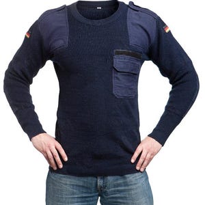 May include: Navy blue sweater with shoulder patches and a chest pocket. The sweater has a ribbed texture and features small German flag patches on the sleeves. The long-sleeved garment is worn with blue jeans.