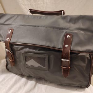 May include: A gray nylon briefcase with brown leather accents. The bag features a flap closure with two leather straps and silver buckles. It has a clear window for a label and a brown leather handle. The bag is designed for carrying documents and other items.