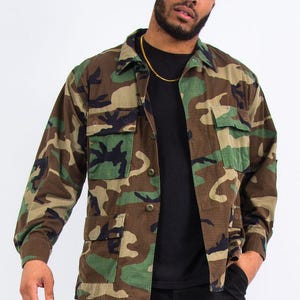 Könnte beinhalten: Eine Tarnjacke in Grün-, Braun- und Schwarztönen. Die Jacke hat eine Knopfleiste, mehrere Taschen und lange Ärmel. Die Person im Bild trägt ein schwarzes Hemd und eine schwarze Hose. Eine goldene Kette ist sichtbar.