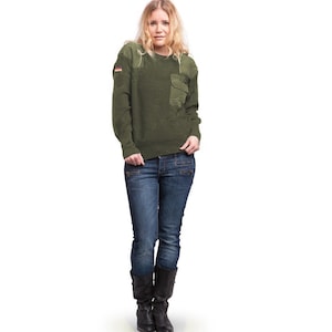 May include: An olive green sweater with a pocket and a German flag patch. The sweater is worn with blue jeans and black boots. The woman is standing against a white backdrop. The sweater is a classic style.