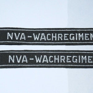 May include: Two black embroidered ribbons with the text "NVA-WACHREGIMENT" in white. The ribbons are rectangular and appear to be made of a woven fabric. The text is centered on each ribbon.