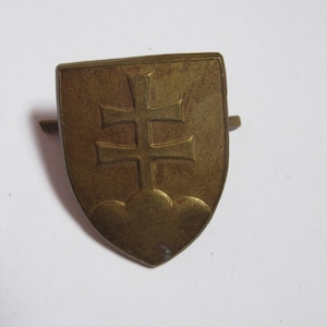Pode incluir: Um alfinete em forma de escudo de cor bronze com um desenho de cruz dupla sobre três nuvens estilizadas. O alfinete tem um design simples e clássico e é provavelmente uma peça vintage ou antiga.