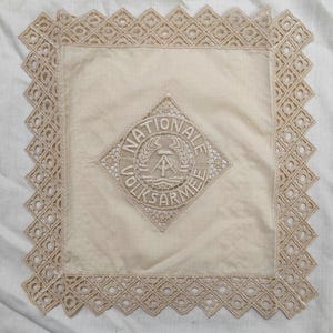 May include: Cream-colored square textile with a decorative border and a central emblem. The emblem features the text "NATIONALE VOLKSARMEE" in a circular design, with a hammer and compass symbol.
