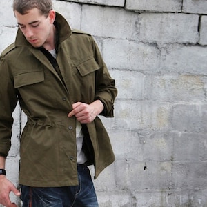 Puede incluir: Chaqueta verde oliva con cierre de botones, dos bolsillos en el pecho y cintura ajustable con cordón. Se lleva sobre una camisa blanca y vaqueros azules. Se ve un reloj de pulsera.