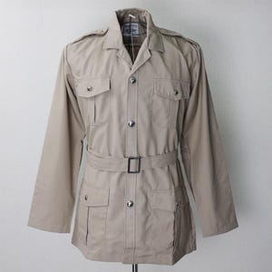以下が含まれることがあります： ベージュ色の長袖ジャケット。襟付きで、ウエストにはベルトが付いています。ジャケットには、ボタン留めのフロントポケットが4つあり、前面にはシルバー色のボタンが付いています。マネキンに着せて展示されています。