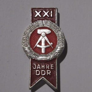 May include: A vintage pin with a red and silver design. The pin features a central emblem with a hammer and compass, surrounded by a wreath. The top banner reads "XXI" and the bottom banner reads "JAHRE DDR".
