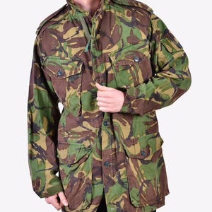 May include: Camouflage jacket in shades of green, brown, and tan. The jacket features multiple pockets, a button-up front, and a high collar. The long-sleeved jacket is made of a durable fabric, suitable for outdoor wear.