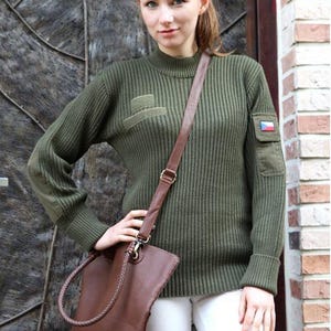 Puede incluir: Un suéter de punto acanalado verde oliva con cuello alto y parches en las mangas. El suéter se combina con pantalones blancos y un bolso de hombro de cuero marrón. Se ve un pequeño parche con la bandera checa.