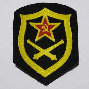 May include: A black patch with a yellow shield design. Inside the shield is a red star with a hammer and sickle, and two crossed yellow cannons. The patch is likely a military or historical emblem.
