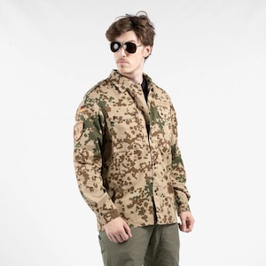 May include: A camouflage-patterned shirt in shades of tan, brown, and green. The shirt features two chest pockets and a patch on the left sleeve. This military-style shirt is part of a clothing collection.