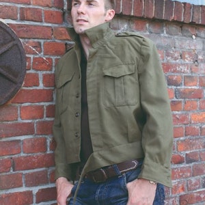 May include: Olive green jacket with four front pockets, button closures, and a collared neckline. The jacket is paired with blue jeans and a brown belt. The model is leaning against a red brick wall.
