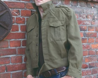 May include: Olive green jacket with four front pockets, button closures, and a collared neckline. The jacket is paired with blue jeans and a brown belt. The model is leaning against a red brick wall.