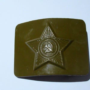 Puede incluir: Hebilla de cinturón verde oliva con un diseño de estrella en relieve y un emblema de martillo y hoz. La hebilla tiene forma rectangular con esquinas redondeadas y un acabado metálico.