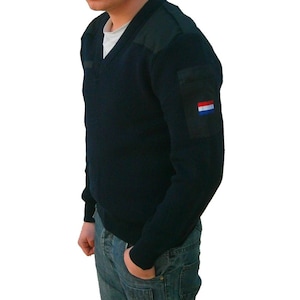 Może przedstawiać: Granatowy sweter z dekoltem w serek, naszywkami na ramionach i małą kieszenią na lewym rękawie. Kieszeń posiada naszywkę z flagą holenderską. Sweter jest noszony na białej koszulce i w połączeniu z niebieskimi dżinsami.