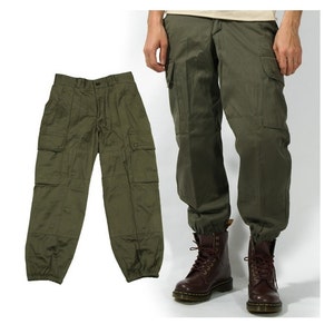 May include: Olive green cargo trousers with multiple pockets and elasticated cuffs. The trousers are shown folded and worn, highlighting their design and fit. The fabric appears to be a durable, lightweight material.