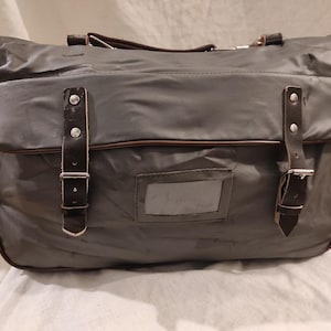 Puede incluir: Un bolso mensajero gris con ribetes y correas de cuero marrón. El bolso tiene forma rectangular con un cierre de solapa asegurado por dos correas de cuero con hebillas metálicas. Una ventana transparente está en la parte delantera. El bolso tiene un asa en la parte superior.