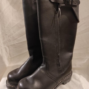 Puede incluir: Un par de botas negras de cuero hasta la rodilla. Las botas tienen una puntera redondeada, una suela gruesa y un detalle de hebilla en la parte superior. El cuero tiene un aspecto texturizado. Botas diseñadas para la durabilidad y protección.