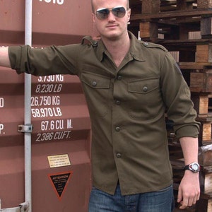 May include: An olive green button-down shirt with two chest pockets and epaulets. The shirt is paired with blue jeans. The person is wearing sunglasses and a watch. The background includes a shipping container with the number 4310.