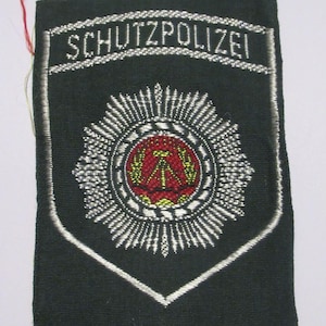 Puede incluir: Un parche negro con la palabra "SCHUTZPOLIZEI" bordada en blanco. Debajo del texto hay un emblema detallado con un centro rojo y amarillo, rodeado por un diseño plateado. El parche tiene un borde blanco.