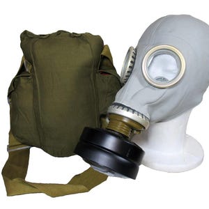 Op de afbeelding: Een grijs gasmasker met heldere lenzen en een zwart filter wordt getoond op een witte mannequinhoofd. Een olijfgroene canvas tas met schouderriem bevindt zich links van het masker.