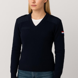 Op de afbeelding: Marineblauwe V-hals trui met schouderstukken en een klein zakje. De trui heeft lange mouwen en een Nederlandse vlag patch op de linkermouw. Een wit t-shirt is zichtbaar eronder. Het model draagt een blauwe jeans.