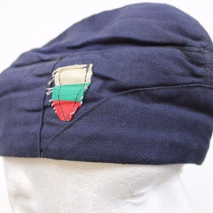 Può includere: Berretto blu navy con una toppa cucita con un disegno bianco, verde e rosso. Il berretto è realizzato in un tessuto morbido e ha una forma arrotondata. La toppa sembra essere una bandiera o un emblema.