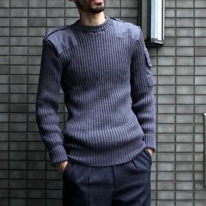 Op de afbeelding: Een marineblauwe ribgebreide trui met versterkte schouderstukken en een klein zakje op de bovenarm. De trui heeft een ronde hals en wordt gedragen met een marineblauwe broek. De algehele stijl is klassiek en praktisch.