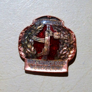 May include: A vintage pin with a red center and silver accents. The pin features text in German and a design of a hammer and other tools. The pin is surrounded by a silver wreath and has a copper-colored border.