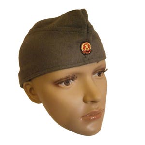 Op de afbeelding: Olijfgroene militaire pet met een rood en goudkleurig embleem. De pet is gemaakt van een viltachtig materiaal en heeft een puntige voorkant. Het embleem is cirkelvormig en heeft een gestileerd ontwerp. De pet wordt getoond op een mannequinhoofd.