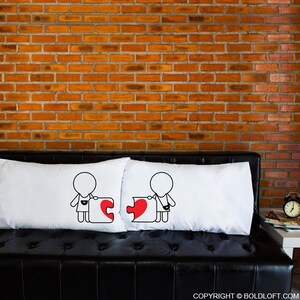 May include: Two white pillowcases with a simple black line drawing of two figures holding puzzle pieces that form a heart. The figures are on a black leather couch in front of a brick wall.