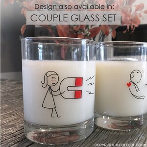 May include: Two clear glass tumblers filled with a white liquid. Each glass has a black and red cartoon design. One glass shows a stick figure with a magnet, the other a stick figure with a heart. Text above reads "COUPLE GLASS SET".