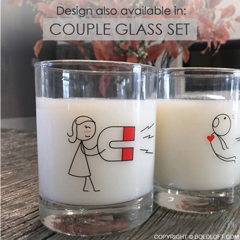 May include: Two clear glass tumblers filled with a white liquid. Each glass has a black and red cartoon design. One glass shows a stick figure with a magnet, the other a stick figure with a heart. Text above reads "COUPLE GLASS SET".