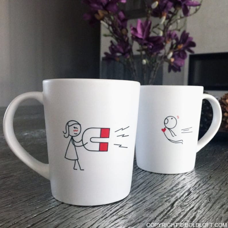 May include: Two white ceramic mugs with black line art illustrations. One mug depicts a person holding a magnet, the other shows a person being pulled towards a heart. The mugs have a curved handle and a simple, modern design.
