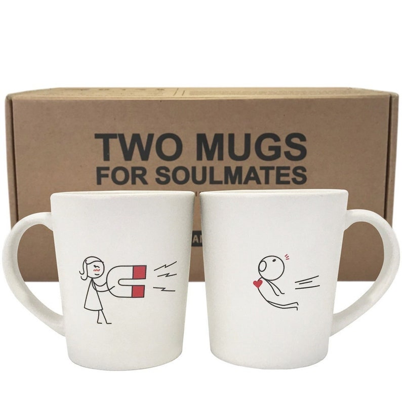 May include: Two white ceramic mugs with black line drawings of stick figures and a magnet. One mug shows a woman with a magnet, the other a figure with a heart. The box behind the mugs reads "TWO MUGS FOR SOULMATES."