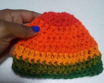May include: A handmade crochet hat in a vibrant color scheme. The hat features bands of green, orange, and red, creating a striped effect. The texture of the crochet stitches is clearly visible, adding to the handmade aesthetic. The hat is held by a hand with blue nail polish.